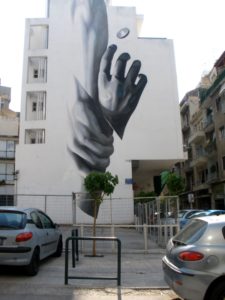 monochrome graffiti on the side of a block of flats in Athens showing a hand reaching down to grab someone else's wrist as if to save them